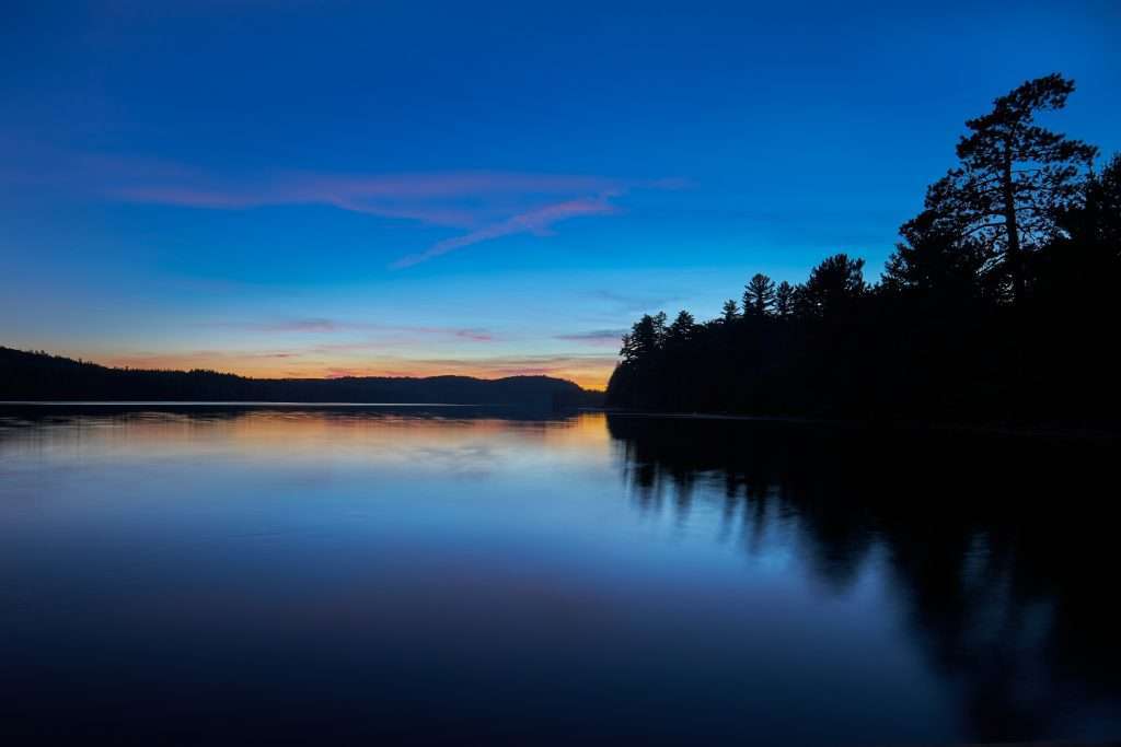 Le soleil vient toujours juste de se coucher, l'horizon est encore dorée alors que le ciel et son reflet sur le lac est d'un bleu roi. La rive, de l'autre côté du lac, trace une rangée d'arbres sous la forme de silhouettes noires. 